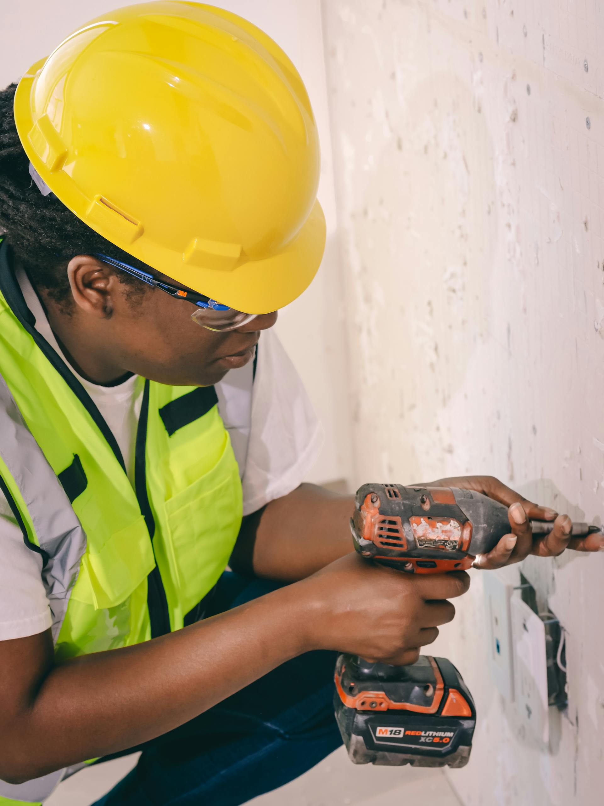Black female construction worker wearing safety gear operates a power drill indoors.